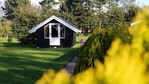 A small, dark wooden cabin with a white door and a triangular roof, surrounded by a well-maintained lawn and trees. - The Comfort of This Cottage Merges With Nature (Holbaek)