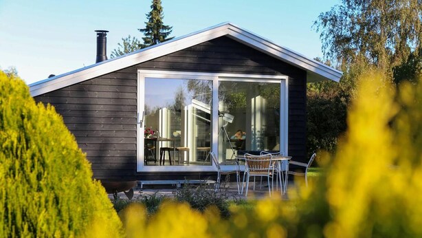 A modern house with a large sliding glass door, a patio with outdoor furniture, and a chimney. - The Comfort of This Cottage Merges With Nature (Holbaek)