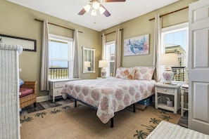 A bedroom with a bed, nightstands, a wicker chair, a window with curtains, and a ceiling fan. - Blue Sky Breeze - 102 Tampico 4 Bedroom Home by RedAwning (Dauphin Island)