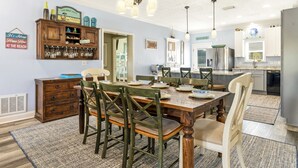 A dining room with a wooden table and chairs, a kitchen in the background, and a wall-mounted cabinet with glassware.