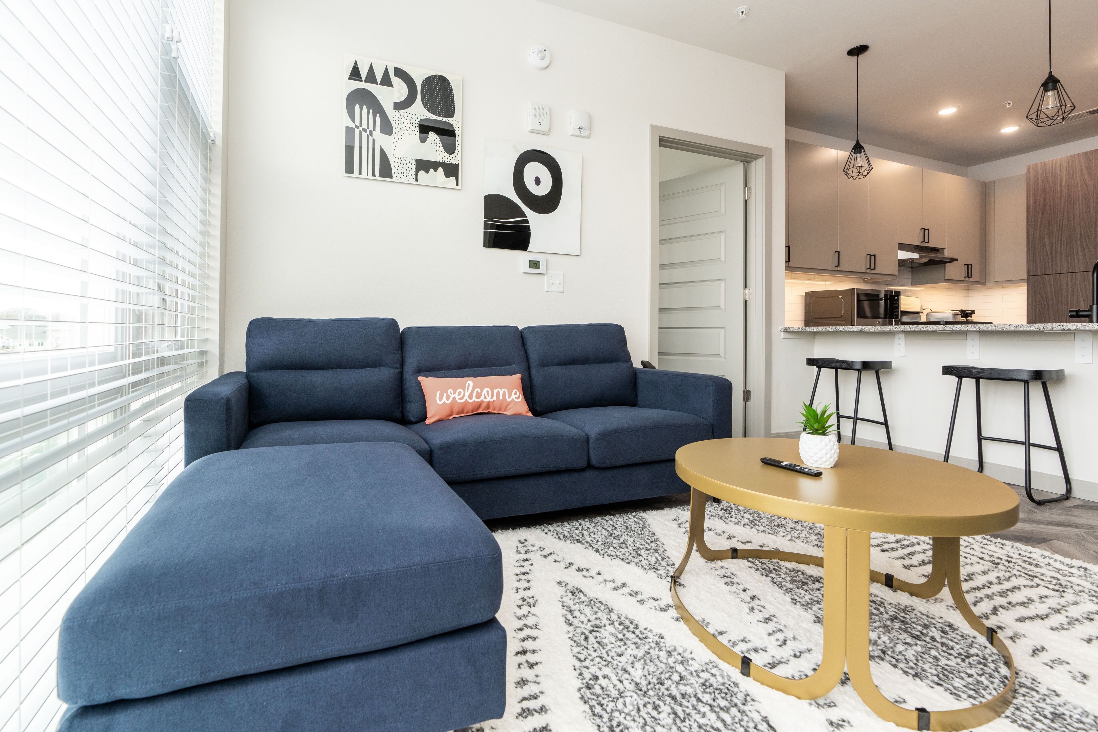 Una sala moderna con un sofá modular, una mesa de centro redonda y una alfombra. Se ve un área de cocina con una encimera y taburetes.