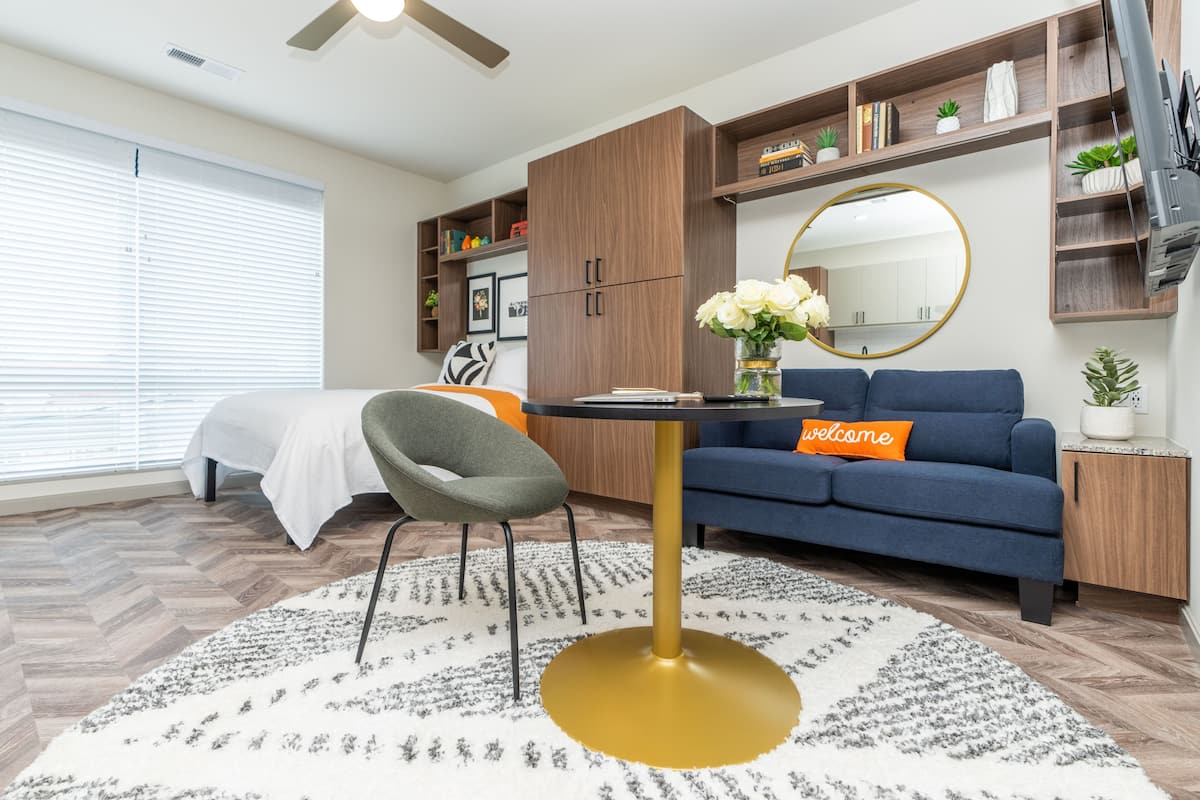 A modern living room with a sofa, a round table, and a chair. There is a bed in the background and a window with blinds.