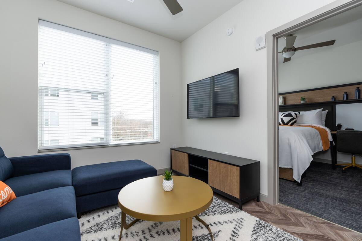 A modern living room with a sofa, a coffee table, a TV, and a bed in the adjacent room.