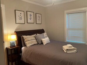 Quarto com cama, almofadas, mesa de cabeceira e luminária, e janela com persianas.