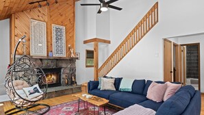 A cozy living room with a stone fireplace, a hanging chair, a blue sofa with patterned cushions, and a wooden staircase.