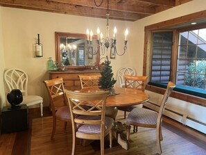 A dining area with a round wooden table, six chairs, a chandelier, a small Christmas tree, and a large window with blinds. - Belleayre Mountain Views (Pine Hill)