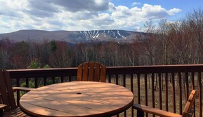 A wooden deck with a round table and chairs, overlooking a forested area and a mountain range.