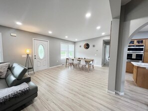 A spacious living area with a dining table and chairs, a sofa, a lamp, and a kitchen area with cabinets and appliances. - Escape to Serenity: 18-Acre Catskill Retreat (Margaretville)