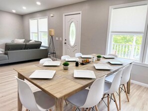 A dining area with a wooden table set for four, surrounded by white chairs, in a room with a grey sofa and a large window. - Escape to Serenity: 18-Acre Catskill Retreat (Margaretville)