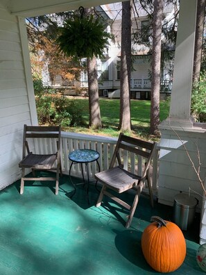 Une véranda avec deux chaises en bois et une petite table ronde, une grosse citrouille orange, et une vue sur un quartier résidentiel avec des arbres et des maisons.
