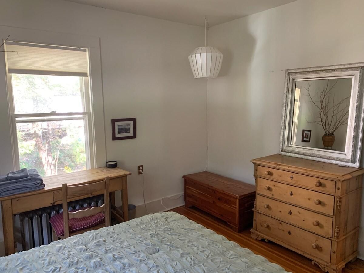 Une chambre à coucher comprenant un lit, une commode, une table de chevet, une fenêtre et un luminaire suspendu.