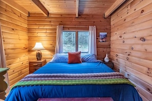 A wooden cabin bedroom with a bed, a nightstand, a lamp, and a window with curtains.