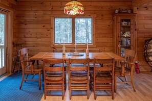 Interior de uma cabana de madeira com uma mesa de jantar, cadeiras, uma janela de vitrais e um armário com louças.