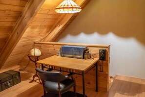 A small, cozy attic room with wooden walls and a sloped ceiling. There is a wooden table with a globe and a bookcase, a black leather chair, and a telephone.