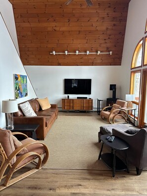 A modern living room with a wooden ceiling, a flat-screen TV, a sofa, a coffee table, and wicker chairs.