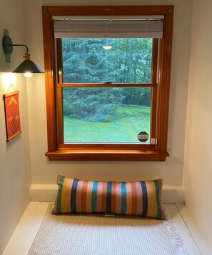A window with a view of a forest, a striped pillow on a bench, and a wall-mounted lamp.