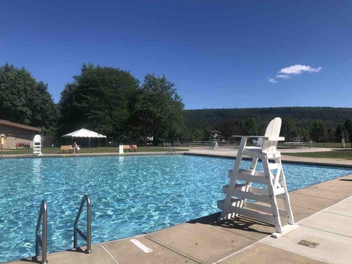 Una piscina con una escalera y una silla de salvavidas, rodeada de árboles y un cielo despejado.