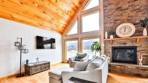 A spacious living room with a high ceiling, wooden beams, a fireplace, a large window, and a flat-screen TV. - Lakeside Catskills Escape • Soak, Sip & Stargaze (Cairo)
