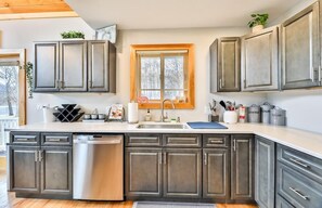 Una cocina con armarios oscuros, un lavavajillas de acero inoxidable, una ventana con persianas y una encimera con varios objetos.