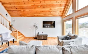 Amplia sala de estar con techo de madera, una escalera, un conjunto de sofás, un televisor de pantalla plana y ventanas grandes.