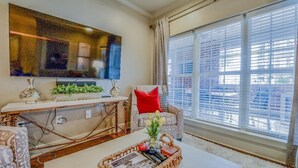 Una sala de estar con un televisor de pantalla plana grande, una mesa de centro con una bandeja, un sofá estampado y una ventana con persianas.