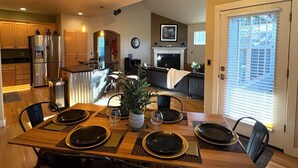 On aperçoit un coin repas avec une table en bois dressée pour quatre personnes, entourée de chaises. Au fond, on distingue une cuisine avec des placards en bois et un réfrigérateur.
