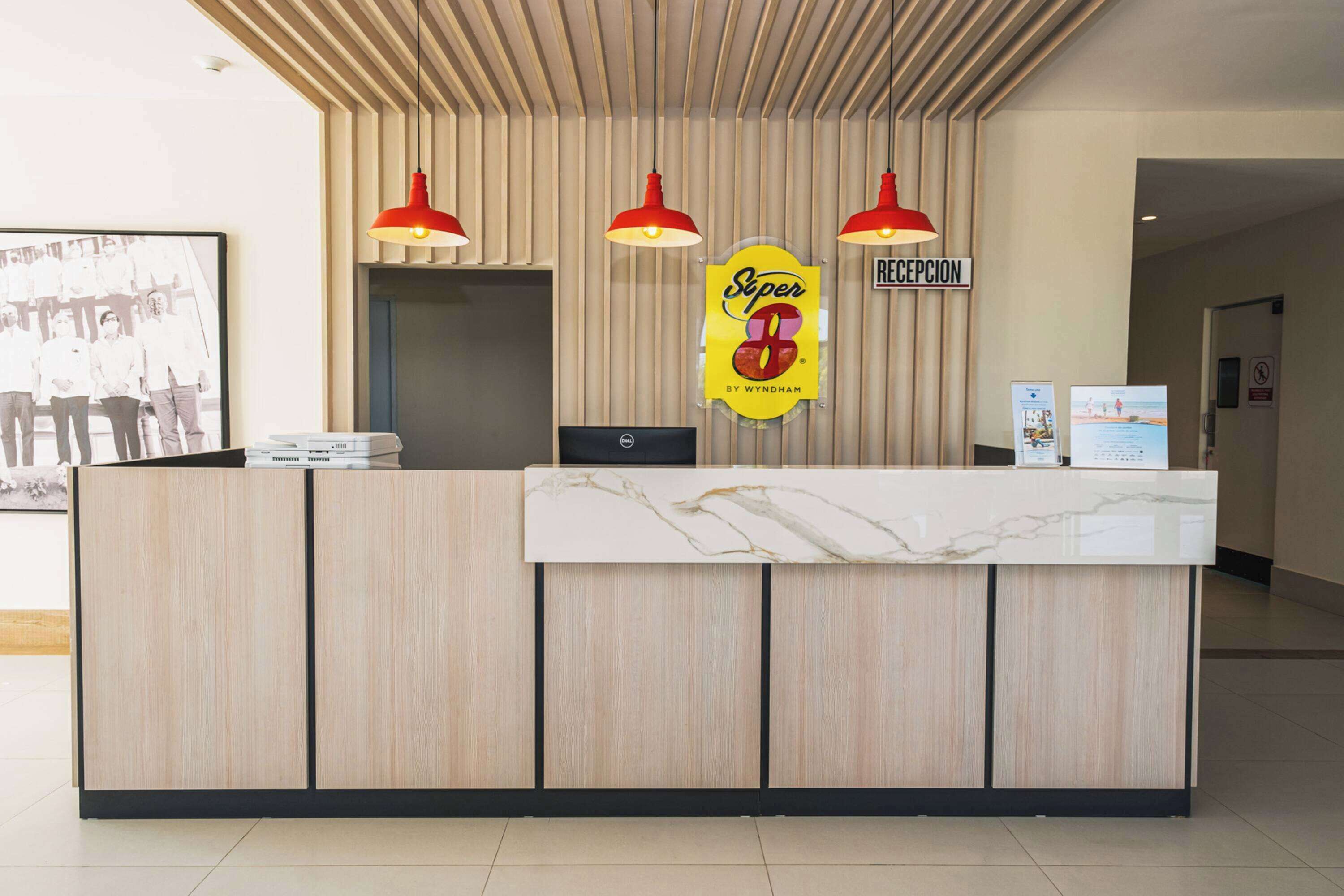 A reception desk with a marble top, wooden paneling, and red pendant lights. A sign for 'Super 8 by Wyndham' and a 'Reception' sign are visible.