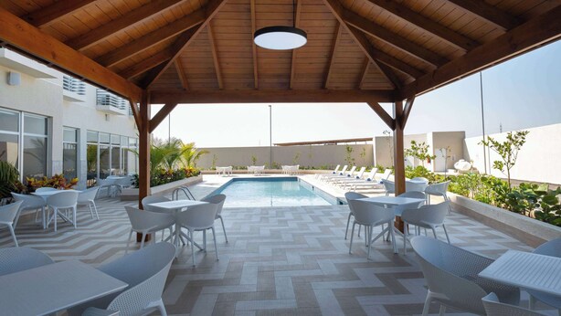 A covered outdoor area with a pool, white chairs, and tables, featuring a wooden ceiling and a tiled floor.