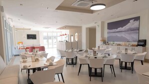 A modern dining area with white tables and chairs, a reception desk, and a large painting on the wall.