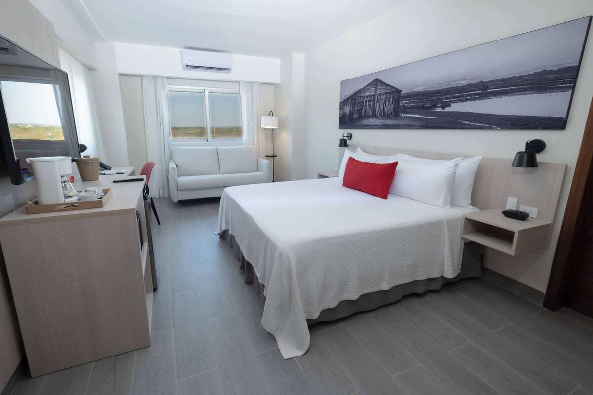 Une chambre d’hôtel avec un lit, un bureau, une chaise, une fenêtre et un tableau au mur.