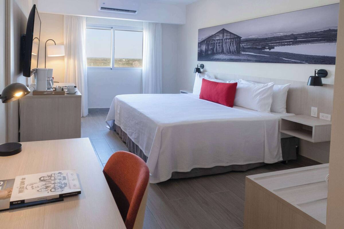 Une chambre d’hôtel avec un lit, un bureau, une chaise, une lampe, un tableau au mur et une fenêtre.