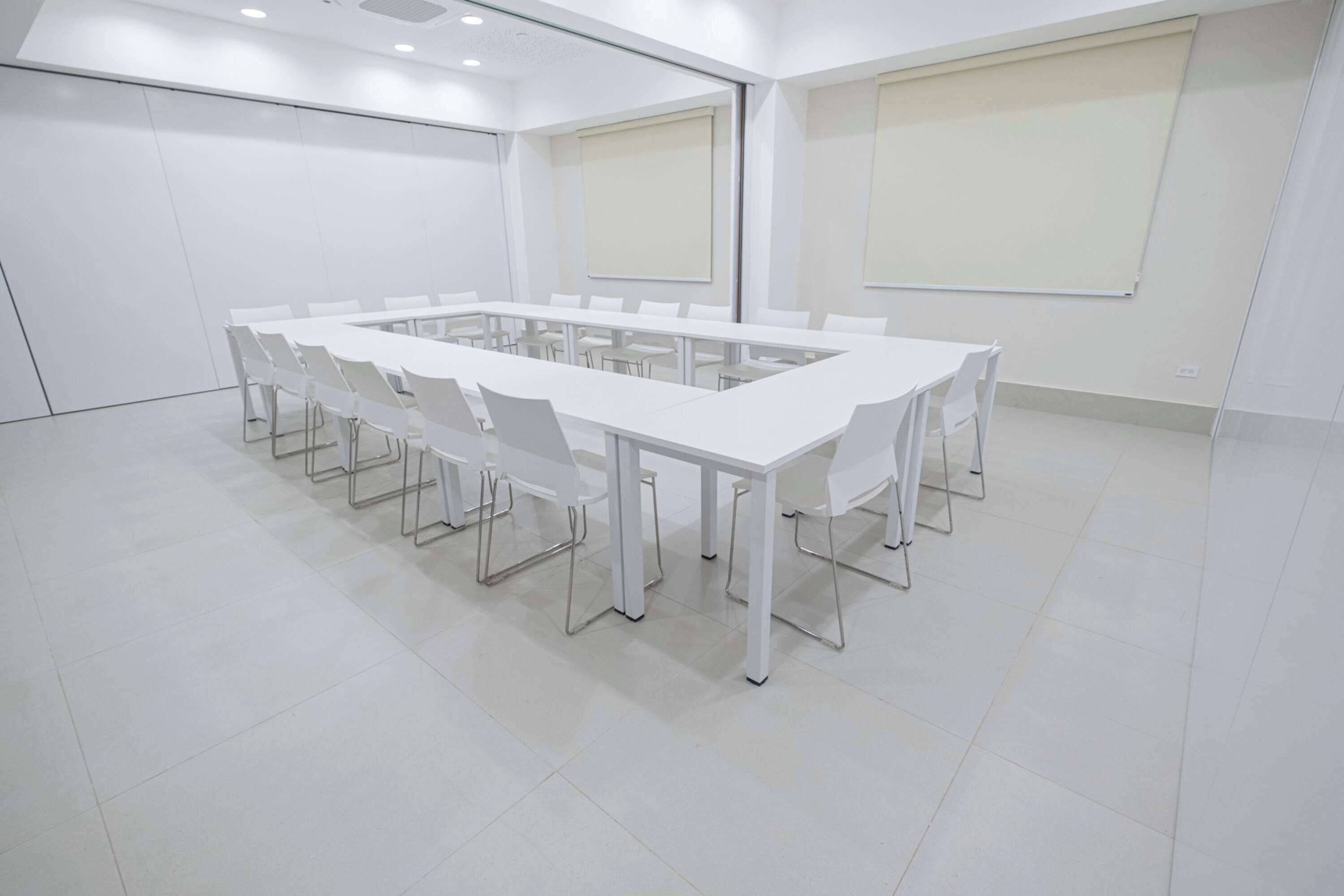 Un salón de conferencias con una larga mesa blanca rodeada de sillas blancas, ventanas amplias y una pantalla de proyector en la pared.