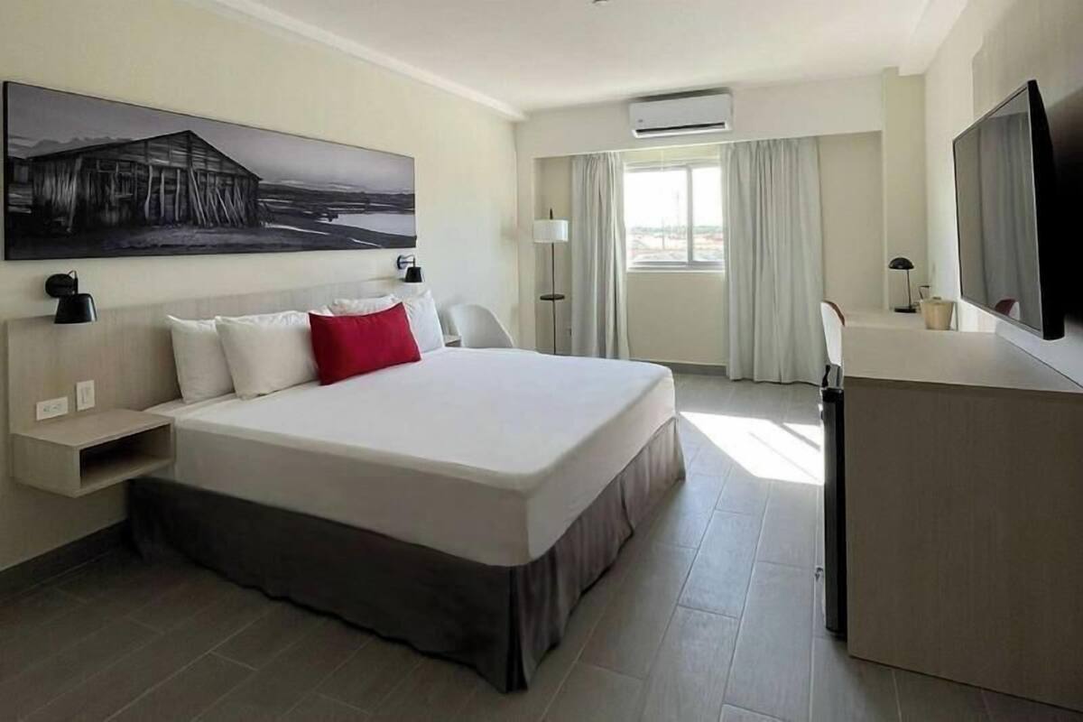 Une chambre d’hôtel avec un grand lit, un bureau, un téléviseur à écran plat, un tableau au mur et une fenêtre avec des rideaux.