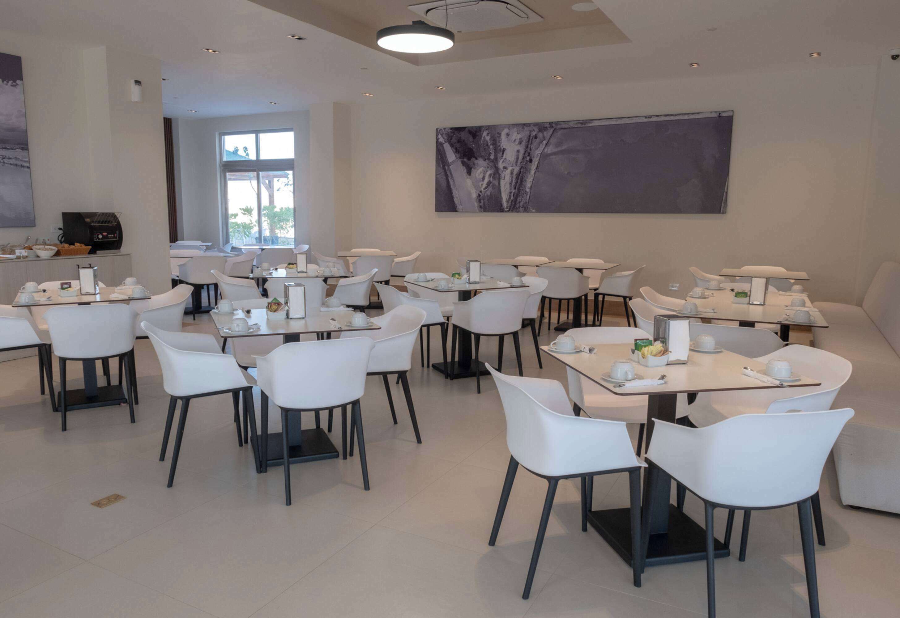 A modern dining area with white chairs and tables, a coffee machine, and a large abstract painting on the wall.