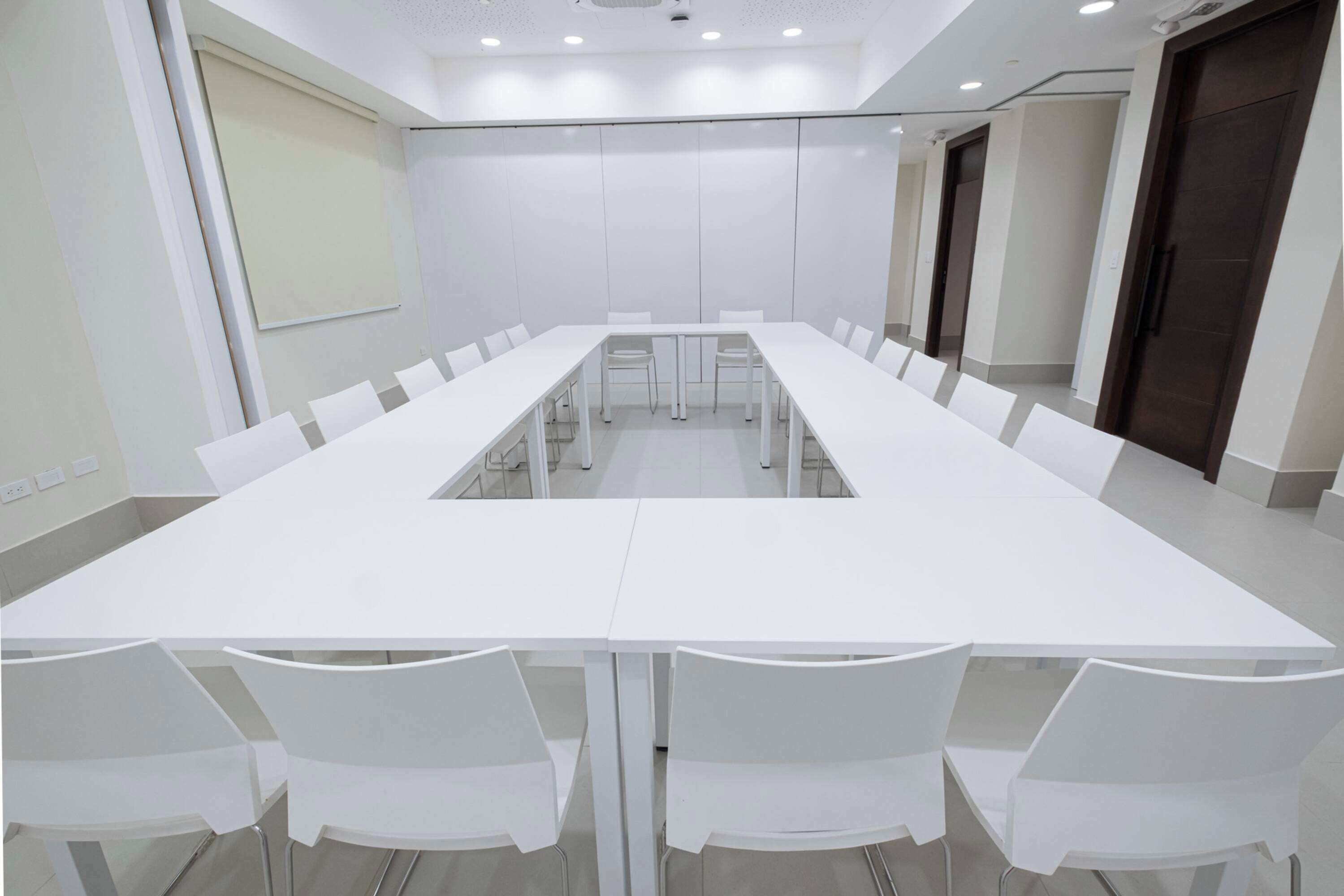 Une salle de conférence avec une longue table blanche et des chaises, des murs blancs et un écran de projection.