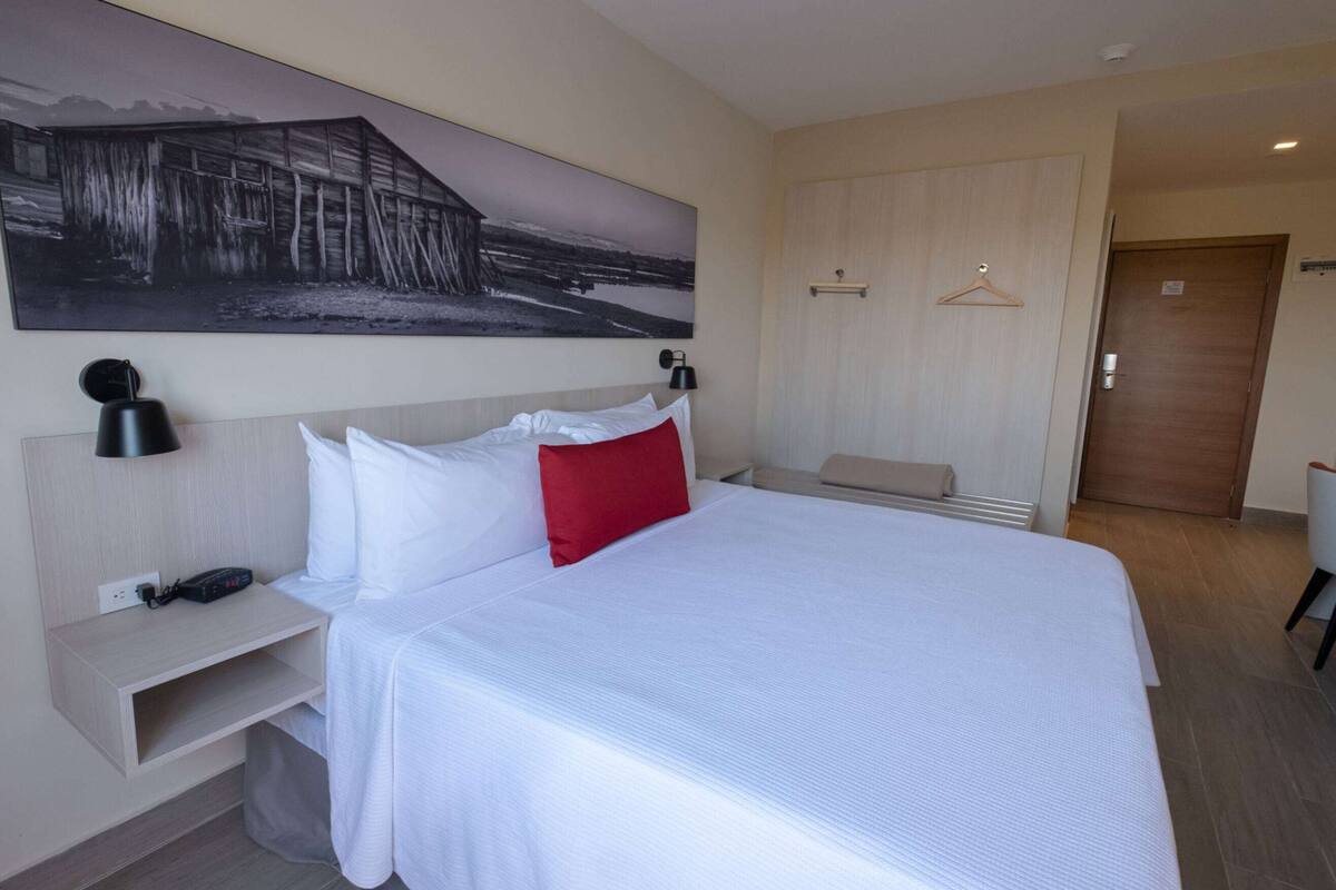 Une chambre d’hôtel avec un grand lit, une table de chevet avec une lampe, un tableau accroché au mur, une porte en bois et un petit coin salon.