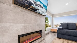 A modern living room with a fireplace, a large flat-screen TV mounted on the wall, and a view of the outdoors through large windows.