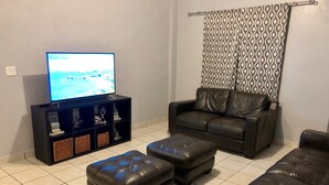 Een woonkamer met een zwarte leren bank, een zwart leren poef, een zwarte entertainmentkast met planken en een televisie op een standaard.