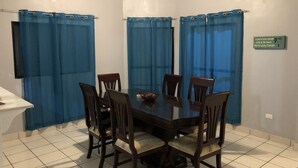 Een eethoek met een donker houten tafel, omringd door zes stoelen, blauwe gordijnen aan de ramen en een bord aan de muur.