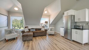 Um loft moderno com sala de estar que inclui um sofá seccional, duas poltronas estampadas e uma cozinha com armários brancos e eletrodomésticos de aço inoxidável.
