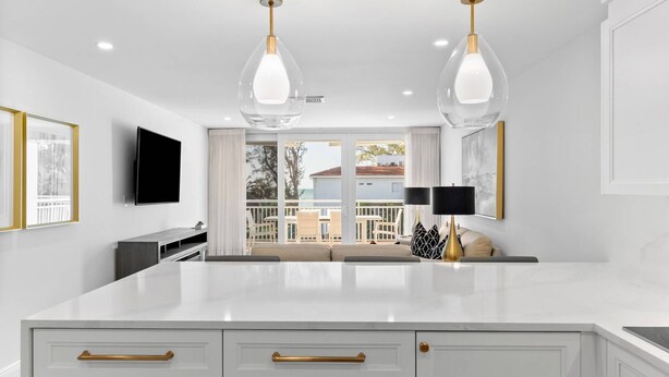 Una cucina moderna con mobili bianchi, una grande isola centrale, luci a sospensione, una TV a schermo piatto e vista su un balcone con mobili da esterno.