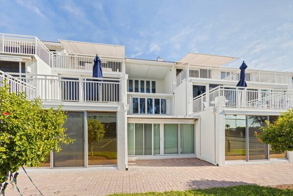 A modern multi-story building with balconies, large sliding glass doors, and a paved entrance area.