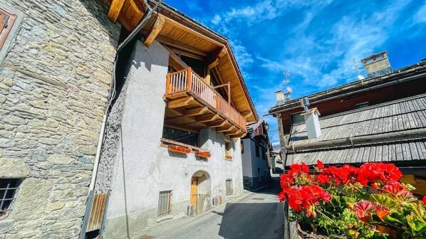 Eine enge Gasse mit Steinhäusern, hölzernen Balkonen und roten Blumen im Vordergrund.