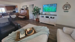 Een woonkamer met een bank, een salontafel met decoratieve objecten, een televisie en een keuken met kasten en een piano.