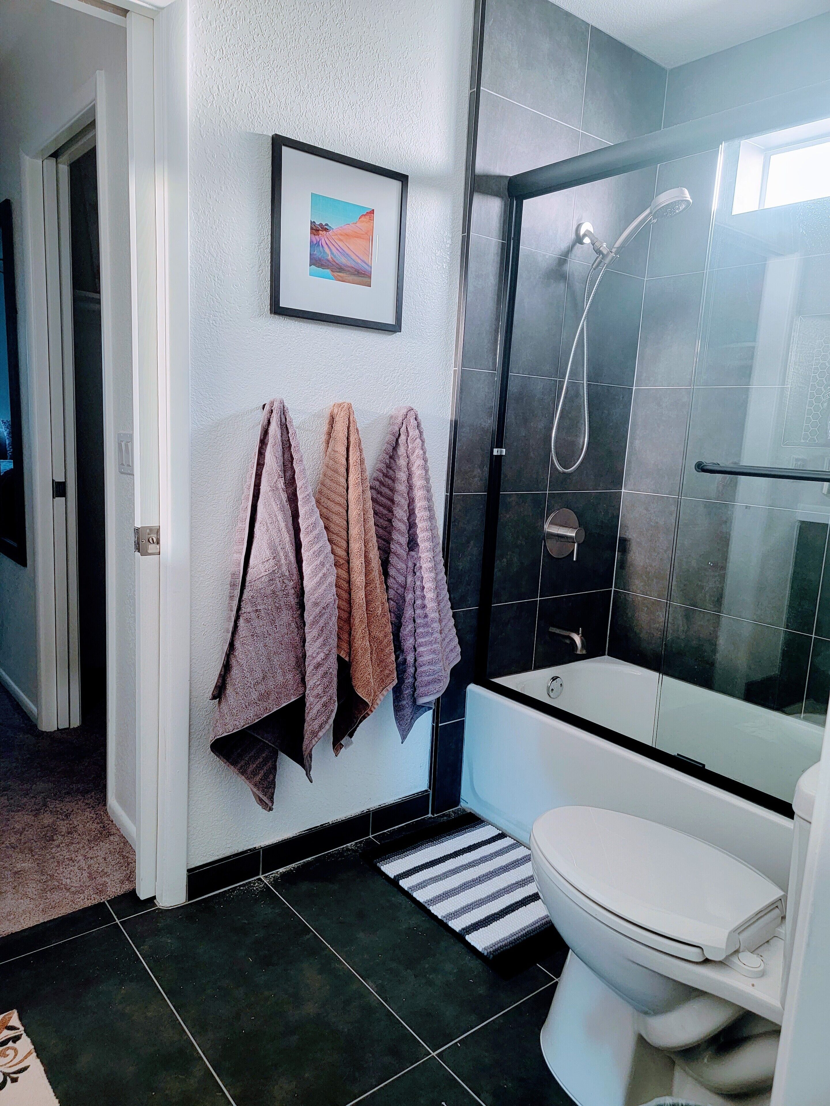 A modern bathroom with a bathtub, toilet, and shower. There are towels hanging on the wall and a framed picture above the sink.