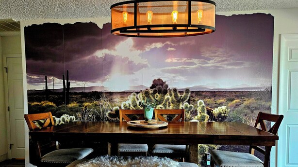 Een eetgedeelte met een grote tafel, stoelen, een kroonluchter en een wanddecoratie van een woestijnlandschap.