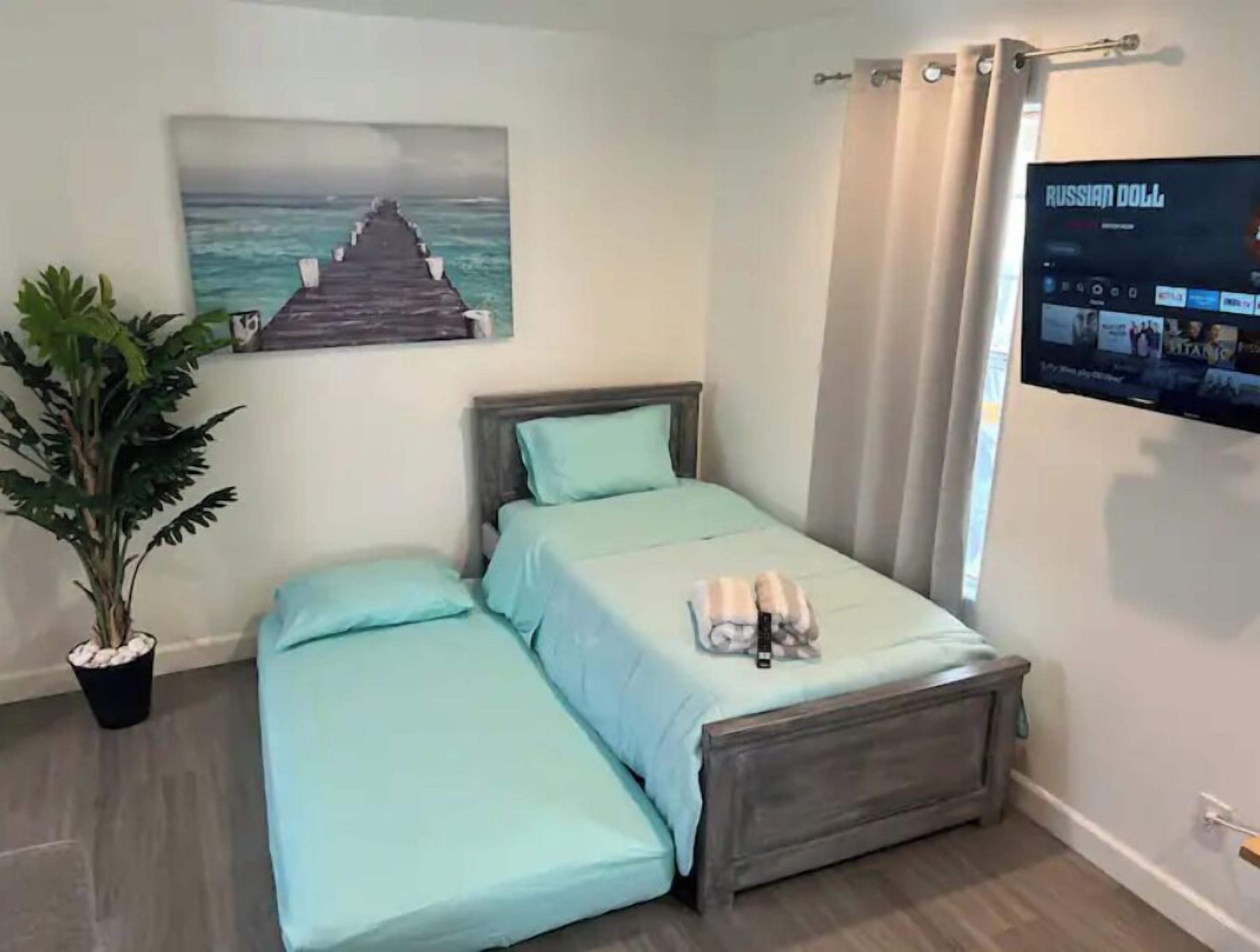 A bedroom with a bed, a potted plant, a painting of a pier, and a TV displaying 'RUSSIAN DOLL'.