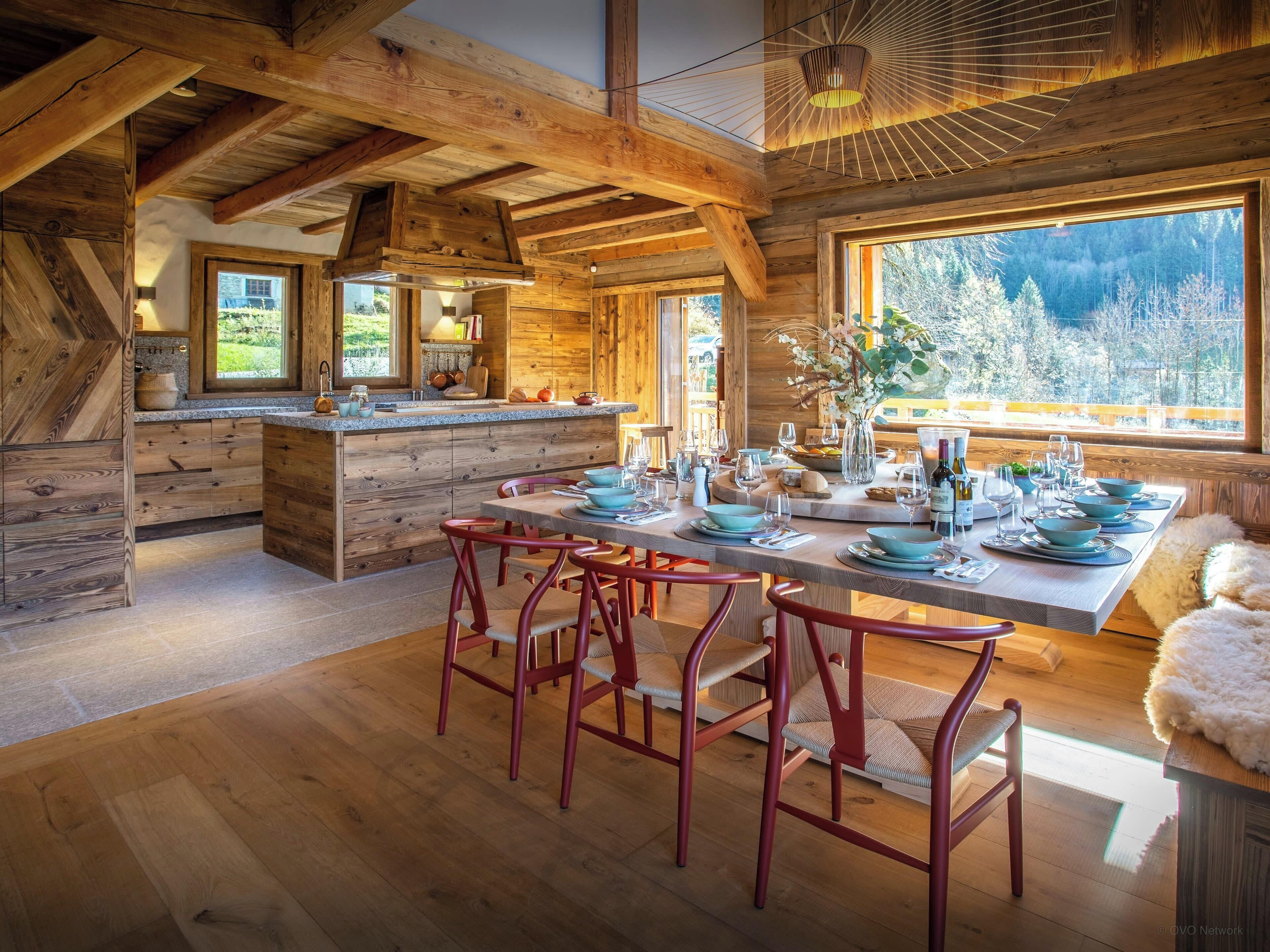 A rustic wooden interior with a dining area, kitchen, and large windows offering a view of the outdoors.