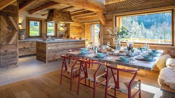 A rustic wooden interior with a dining area, kitchen, and large windows offering a view of the outdoors.