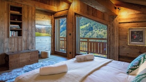 A wooden cabin bedroom with a bed, a nightstand, a bookshelf, and a large window offering a mountain view.